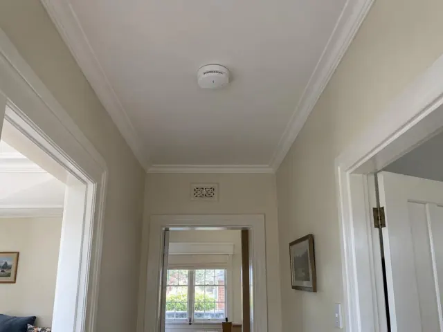 Ceiling-mounted smoke alarm in a hallway with cream walls, white trim and open doorways in Ryde